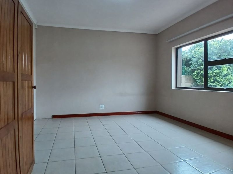 A clean, empty bedroom featuring neutral wall paint, ceramic floor tiles, and built-in wooden cupboards with natural light from the window.