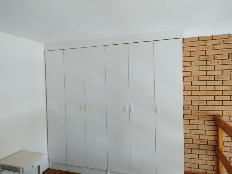 A bedroom featuring extensive built-in white cupboards, an exposed brick feature wall, and warm wooden flooring.