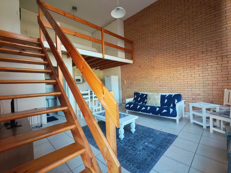 Open-plan living area featuring a double-volume ceiling, exposed brick feature wall, and a wooden staircase leading to a mezzanine level.