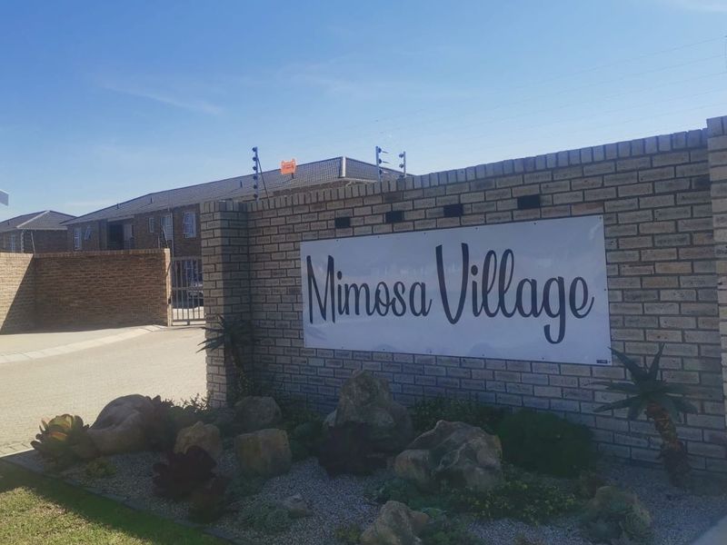 Entrance view of Mimosa Village townhouse complex featuring a brick boundary wall, electric fencing, and a paved driveway.