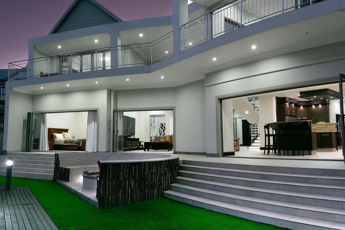 A modern double-story rear elevation featuring a spacious tiled patio, built-in fire pit, and seamless indoor-outdoor flow through large stacking glass doors.