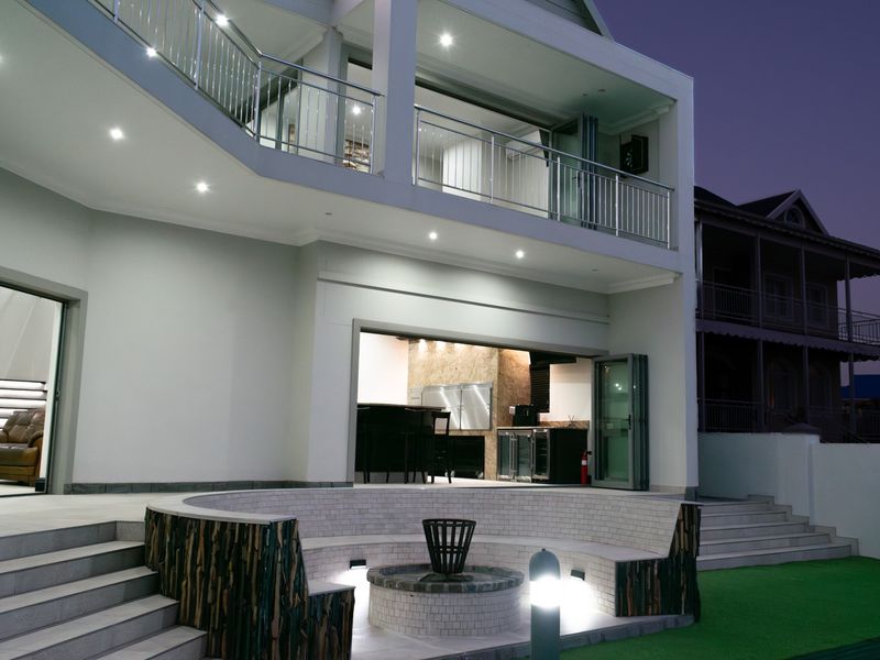A modern rear exterior featuring a multi-level patio, a built-in circular fire pit with stone seating, and a balcony with stainless steel railings.