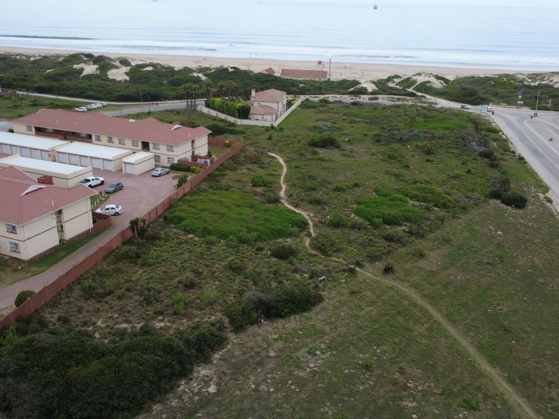 Aerial view of a vacant residential plot situated near the coastline, adjacent to an existing residential complex and main road.