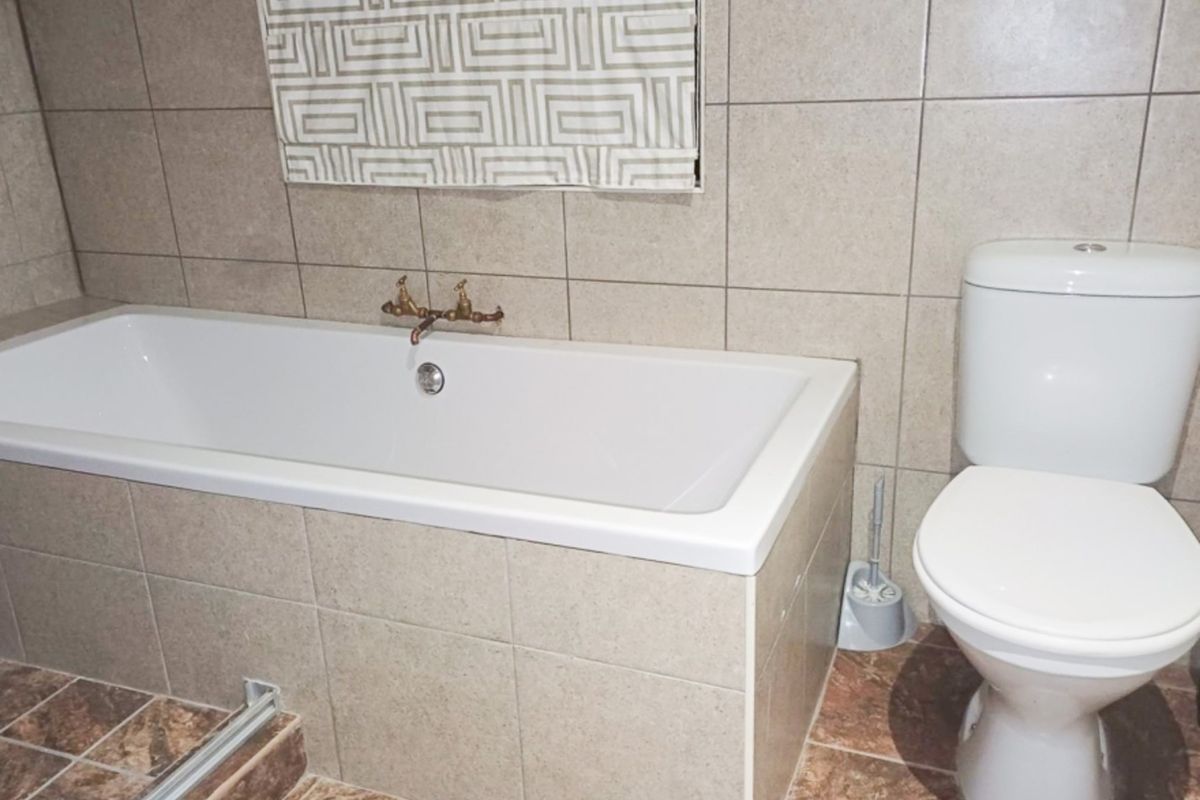 A functional bathroom featuring a built-in bathtub with brass fixtures, a close-coupled toilet, and neutral ceramic wall and floor tiling.