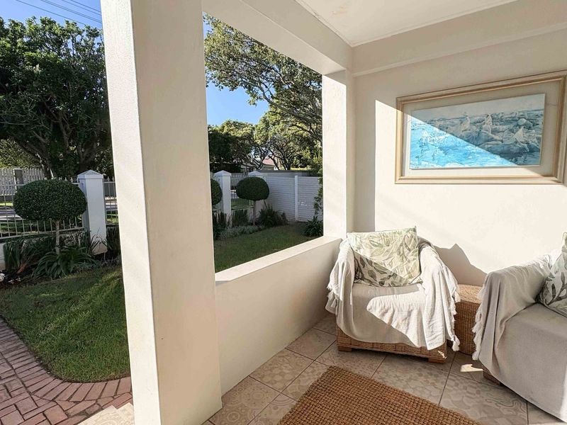 A covered patio area featuring tiled flooring and concrete pillars, overlooking a well-maintained front garden and paved driveway.