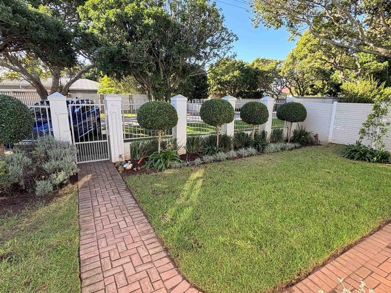 A well-maintained front garden featuring a brick paved walkway, manicured topiary trees, and secure perimeter walling with a metal pedestrian gate.
