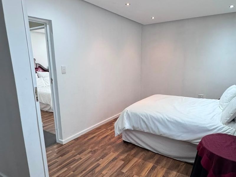 A clean, neutral bedroom featuring modern laminate flooring and recessed downlighting, with an adjoining room visible through the doorway.