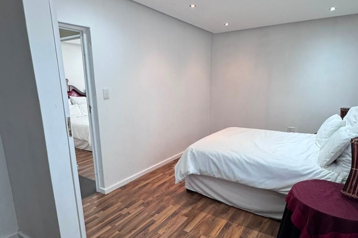 A clean, neutral bedroom featuring modern laminate flooring and recessed downlighting, with an adjoining room visible through the doorway.
