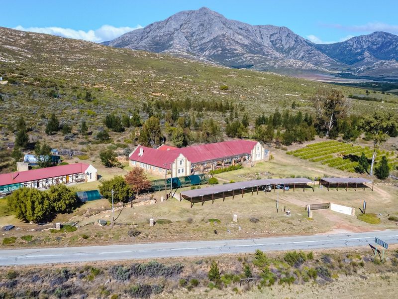 Aerial view of a rural guest house property featuring multiple buildings, extensive grounds, and covered parking structures set against a mountain backdrop.