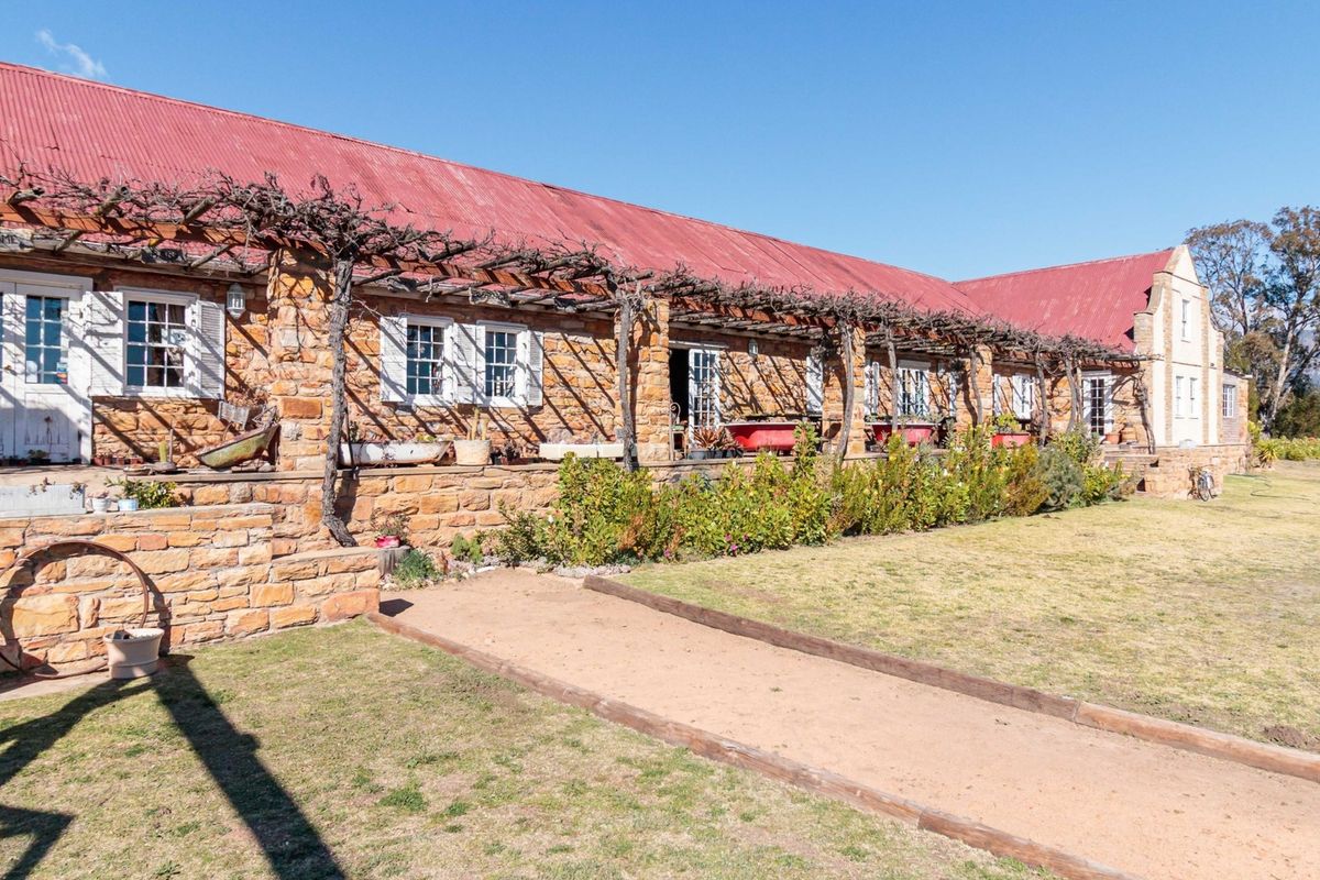 A rustic stone-clad guest house exterior featuring a long timber pergola, corrugated metal roof, and multiple french door access points overlooking the garden.