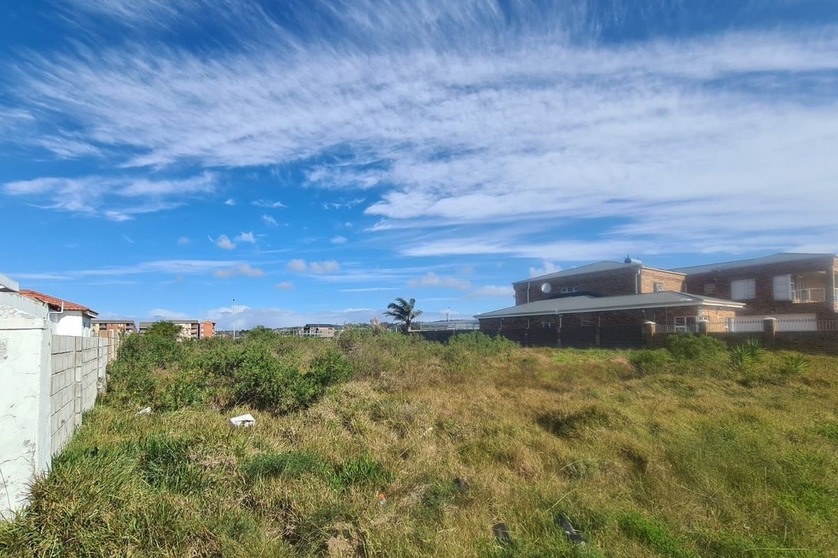 A view of a vacant residential plot featuring overgrown grass and a boundary wall, situated in a developed neighborhood.