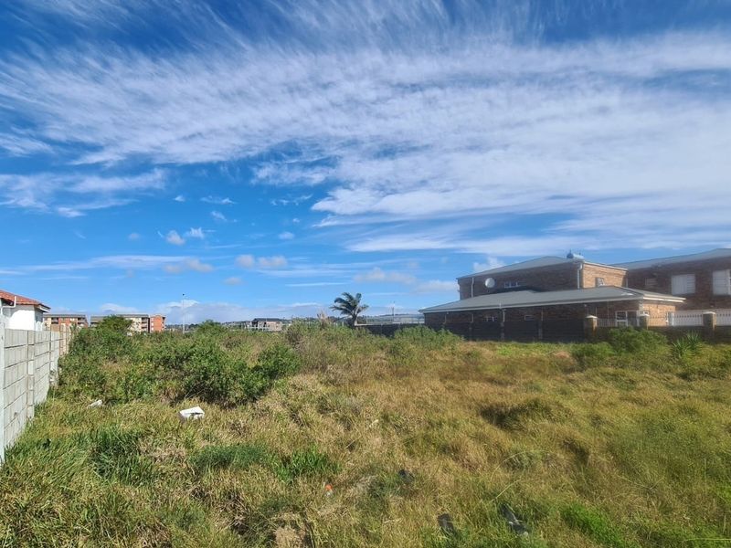 A view of a vacant residential plot featuring overgrown grass and a boundary wall, situated in a developed neighborhood.