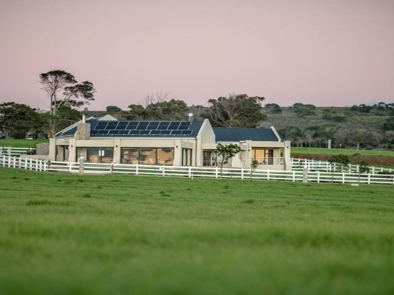 A well-maintained residential property featuring a metal roof with integrated solar panels, a stone chimney, and a white picket fence set in a rural landscape.