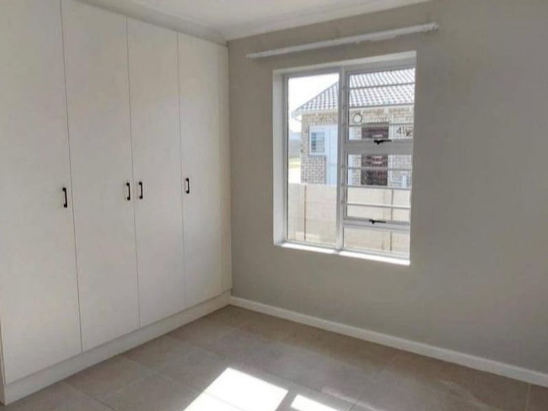 A bright, empty bedroom featuring neutral tiled flooring, built-in cupboards, and a window with security bars.