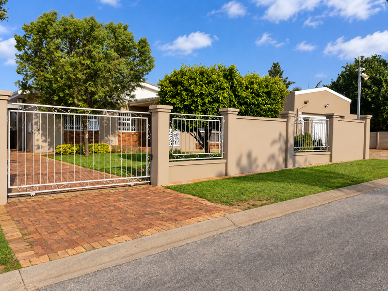 Front view of the property featuring a secure wrought iron gate, perimeter wall, and a well-maintained brick paved driveway and garden area.
