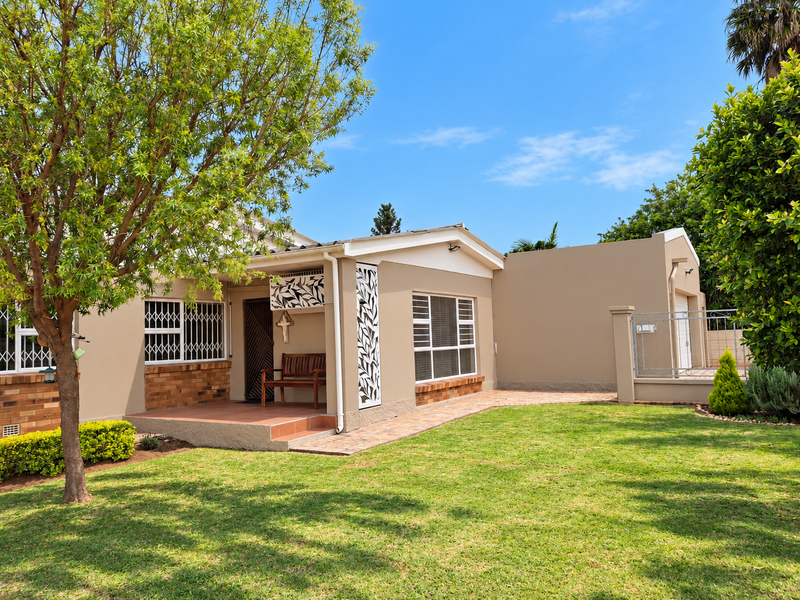 A well-maintained single-story home featuring a tiled front patio, brick accents, secure metal gates, and a neat, landscaped garden with a paved walkway.