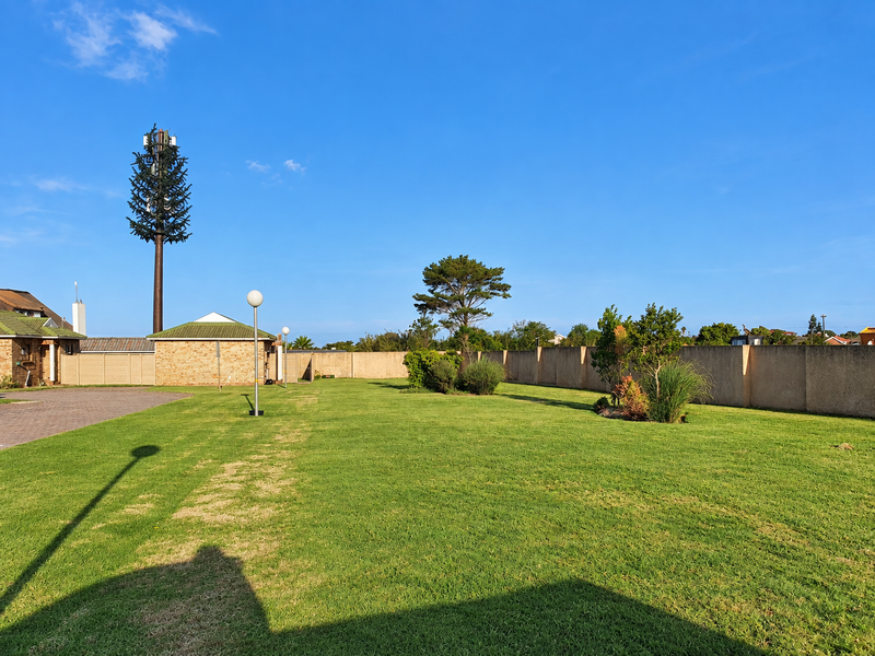 A spacious, well-maintained lawn area featuring a perimeter wall, paved driveway access, and exterior lighting, with a prominent telecommunications tower in the background.