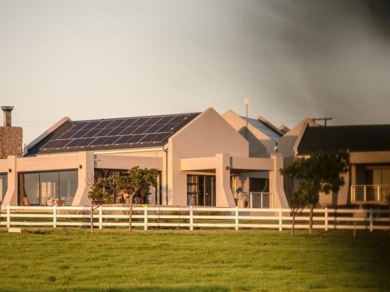 A modern residential property featuring solar panels, a stone chimney, and a covered patio, enclosed by a white picket fence on a grassy lot.