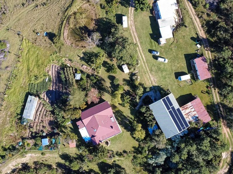 Aerial view of a farm property featuring multiple buildings, solar panel installations, cultivated garden plots, a greenhouse, and surrounding rural landscape.