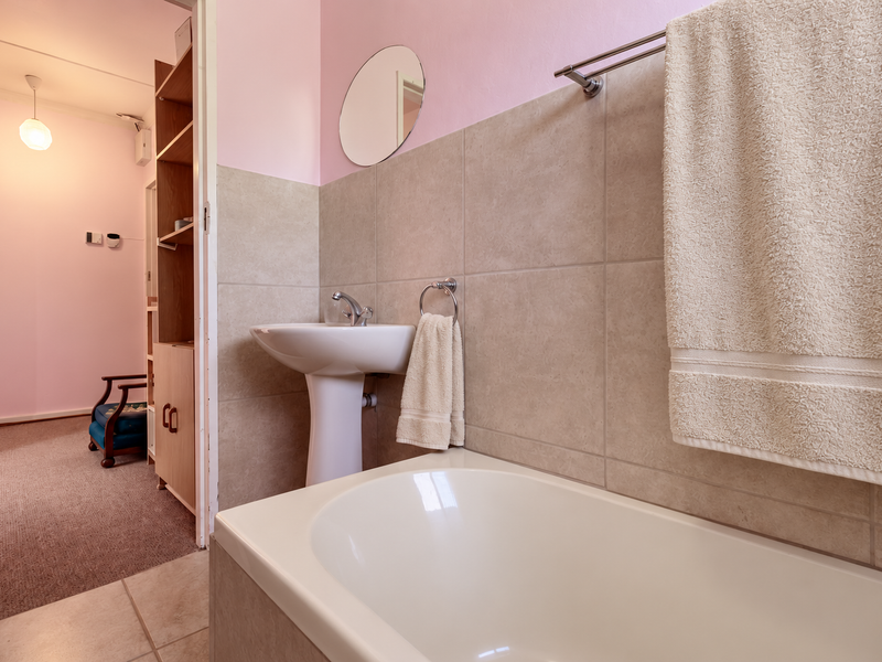 A functional bathroom featuring a pedestal basin, a built-in bathtub with tiled surround, and neutral wall tiling, adjacent to a carpeted living area.