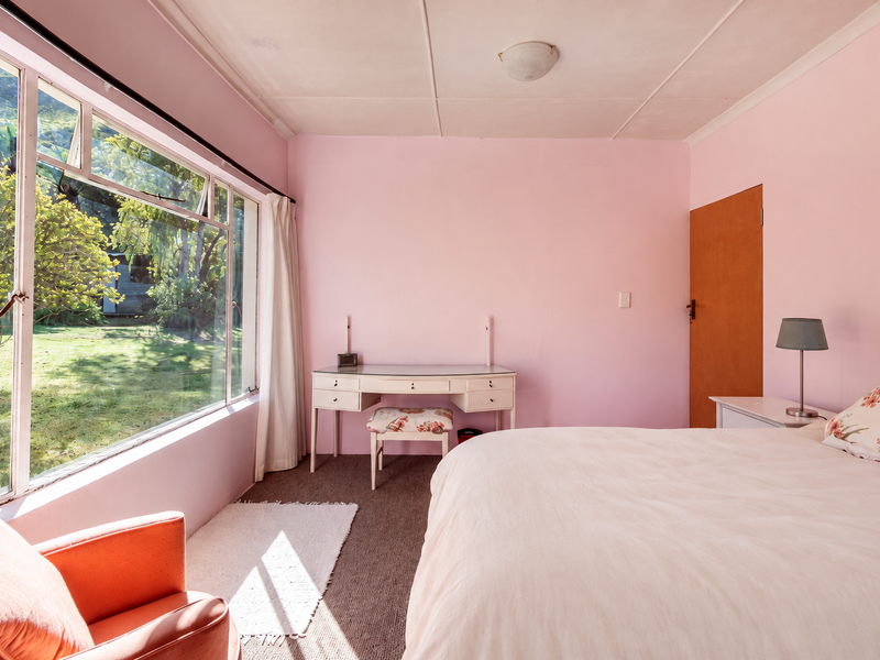 A bedroom featuring pink walls, large windows with garden views, and carpeted flooring, offering a bright space with potential for modernization.