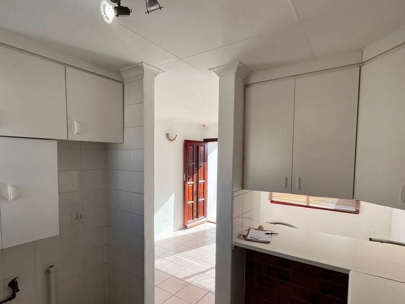 A functional kitchen space featuring white built-in cabinetry, tiled splashbacks, and a clean countertop, leading through to an adjacent living area.