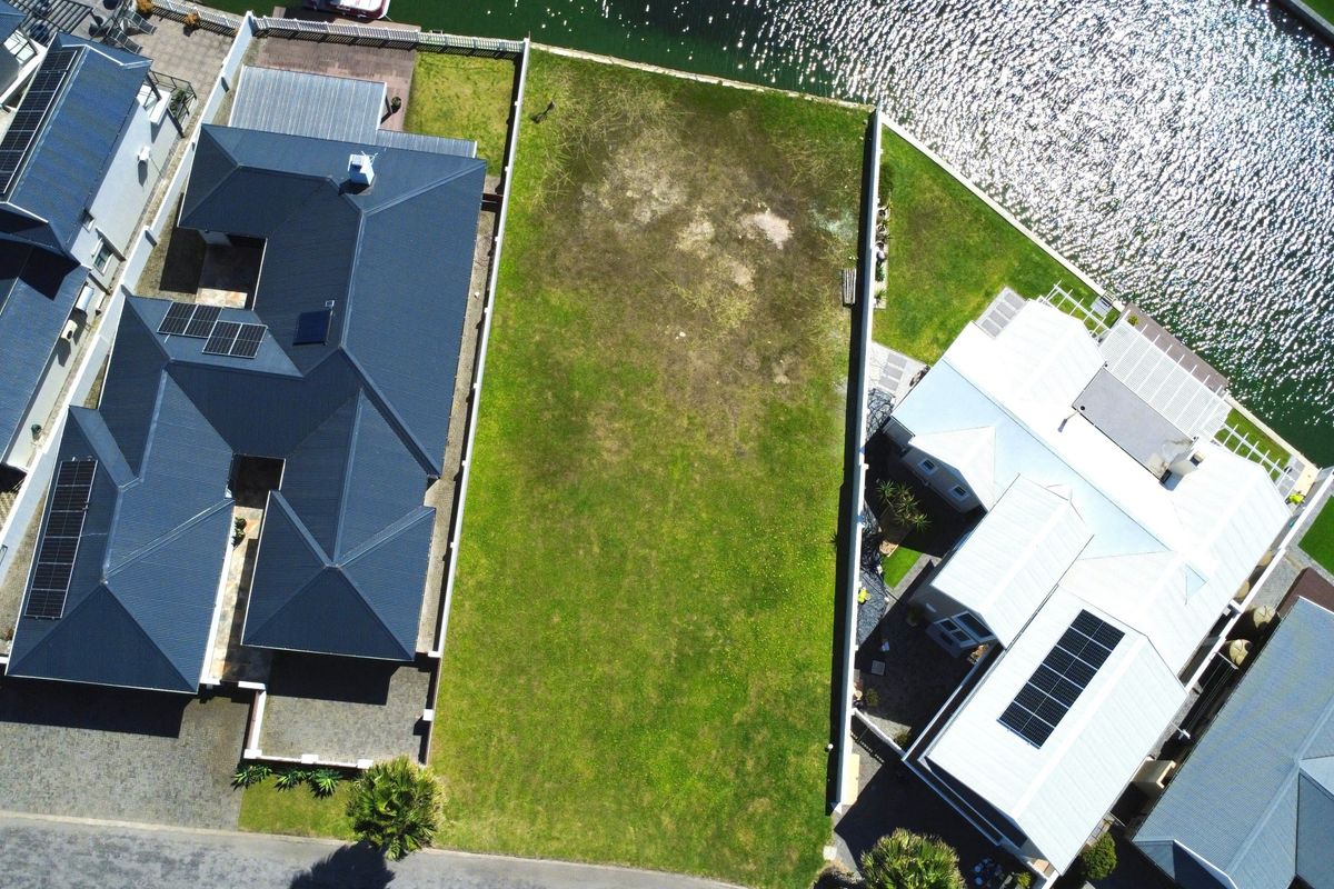Aerial view of a vacant residential plot situated between two established homes, featuring direct waterfront access and clear boundary fencing.