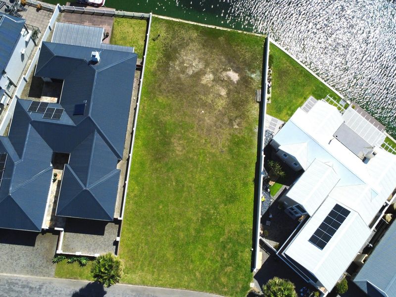 Aerial view of a vacant residential plot situated between two established homes, featuring direct waterfront access and clear boundary fencing.