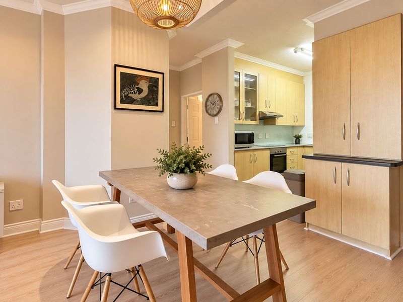 An open-plan dining and kitchen area featuring light wood-toned cabinetry, modern laminate flooring, and integrated kitchen appliances in a well-maintained apartment setting.