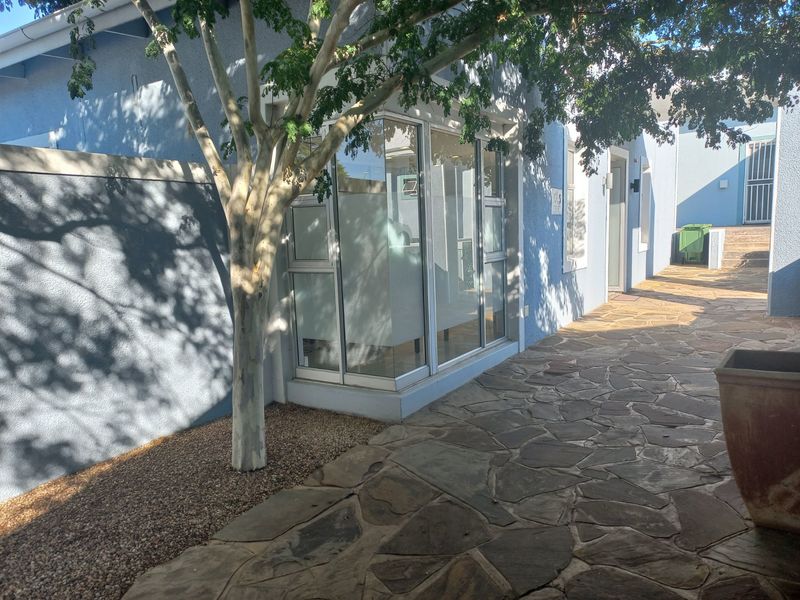 A paved exterior walkway leading along the side of the office building, featuring stone flooring and aluminium-framed windows.