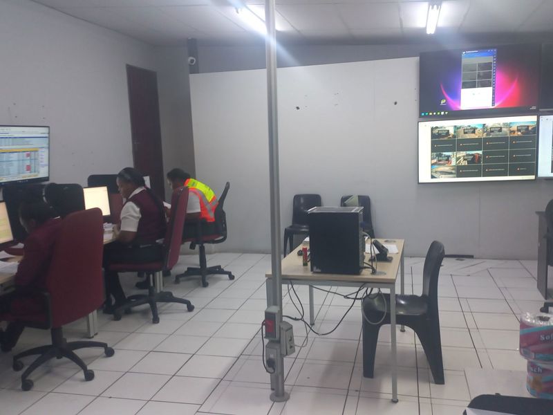 A functional office workspace featuring multiple wall-mounted monitors, tiled flooring, and overhead fluorescent lighting, currently set up as a monitoring or control room.