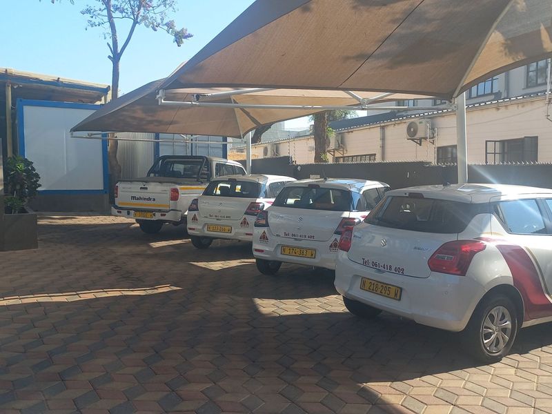 A paved outdoor parking area featuring a large shade sail structure and access to the building via concrete stairs.