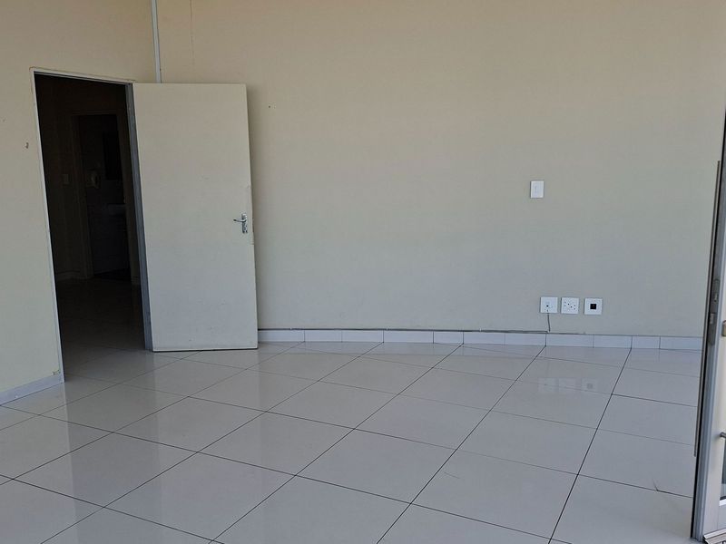 A bright, empty office space featuring neutral tiled flooring, clean walls, and a glass sliding door providing access to an outdoor area.