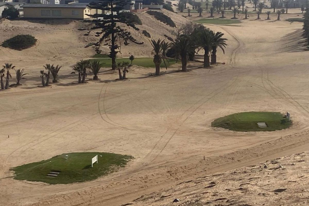 An elevated aerial view of a residential golf course landscape featuring sandy terrain, manicured green patches, and scattered palm trees.