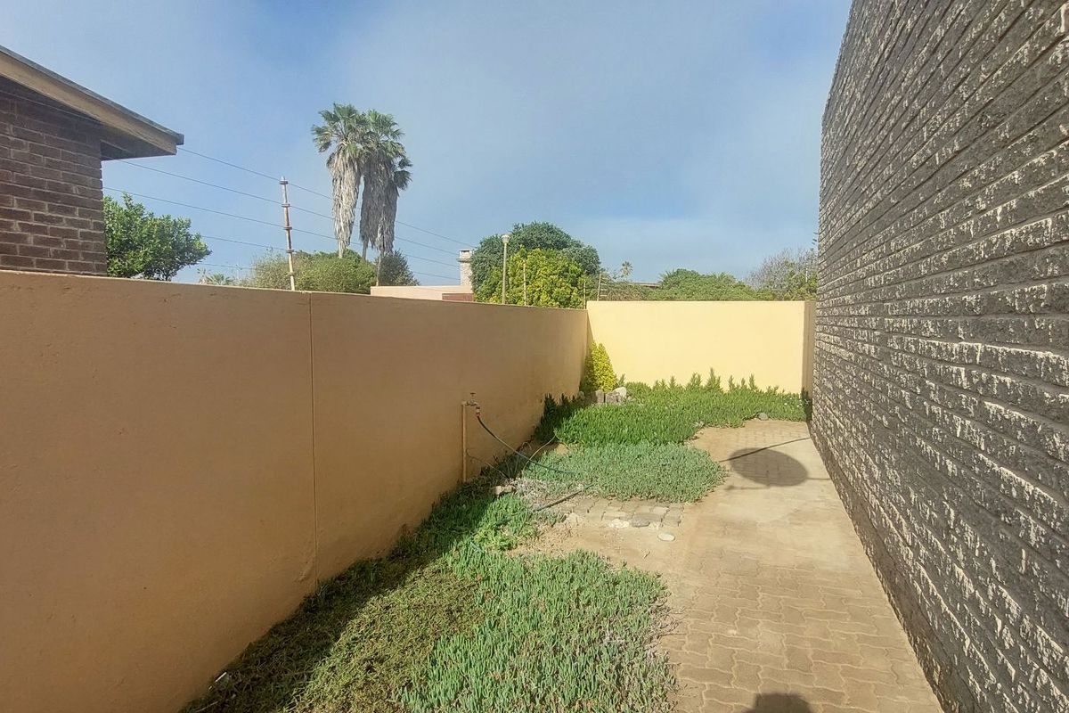 A side exterior area featuring a paved walkway, boundary wall, and established ground cover vegetation.