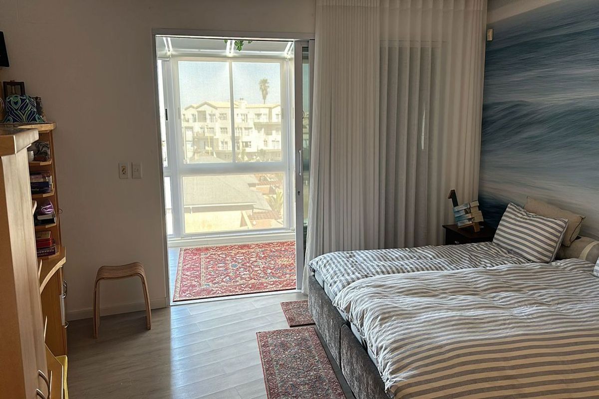 A bright bedroom featuring light wood-look flooring, a large sliding glass door leading to a balcony, and floor-to-ceiling curtains.