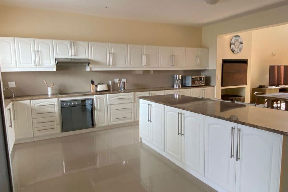 Spacious kitchen featuring white cabinetry, stone countertops, a central island, and integrated oven and extractor fan, flowing into an adjacent living area.
