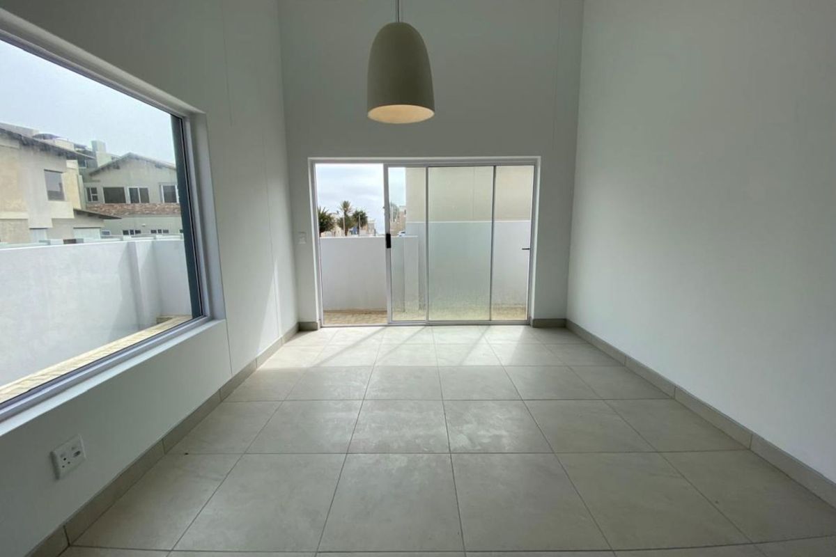 A bright, empty living room featuring large windows, sliding glass doors leading to an outdoor area, and neutral floor tiling.
