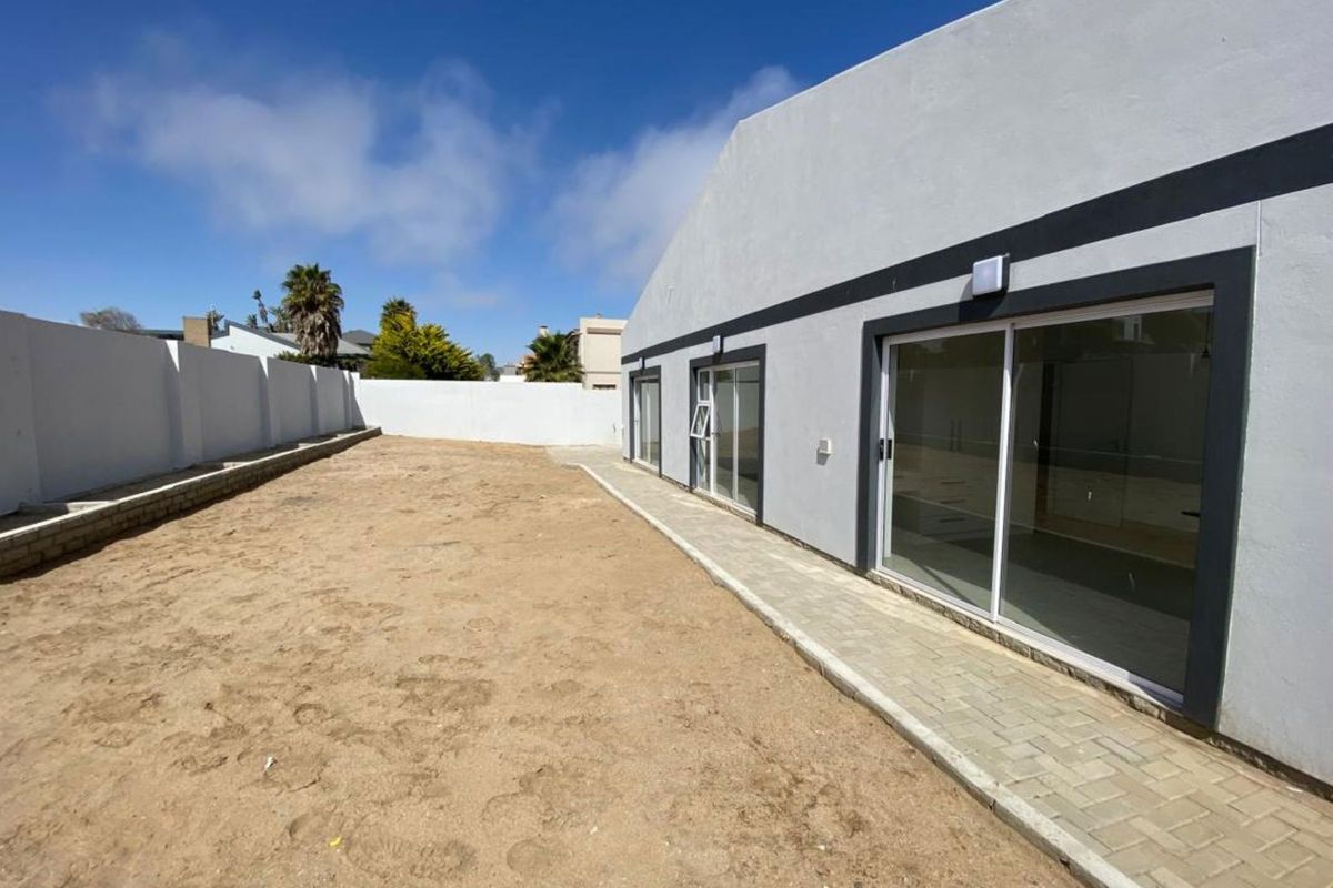 Rear view of the property featuring a paved walkway, sliding glass doors, and a large, undeveloped sandy backyard enclosed by perimeter walls.