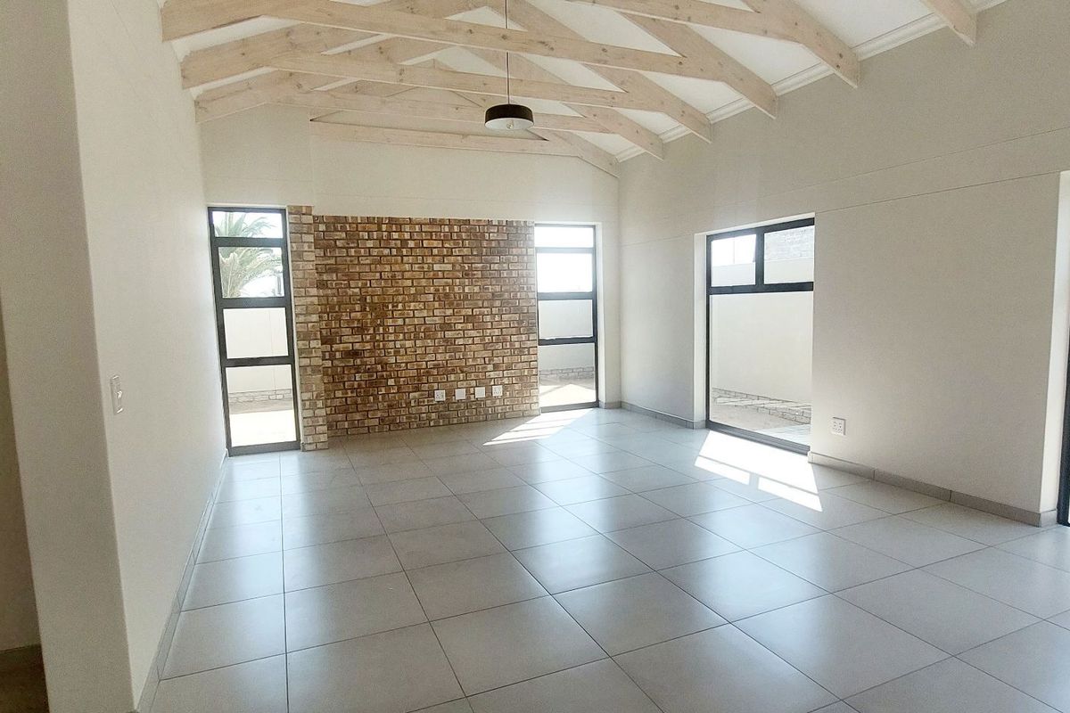 Spacious living area featuring exposed timber roof trusses, a decorative face-brick feature wall, large windows, and neutral floor tiling.