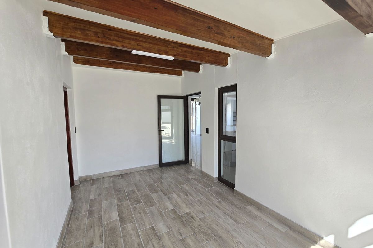 A bright, empty living space featuring exposed timber ceiling beams and modern wood-look tiled flooring with glass-paneled doors.