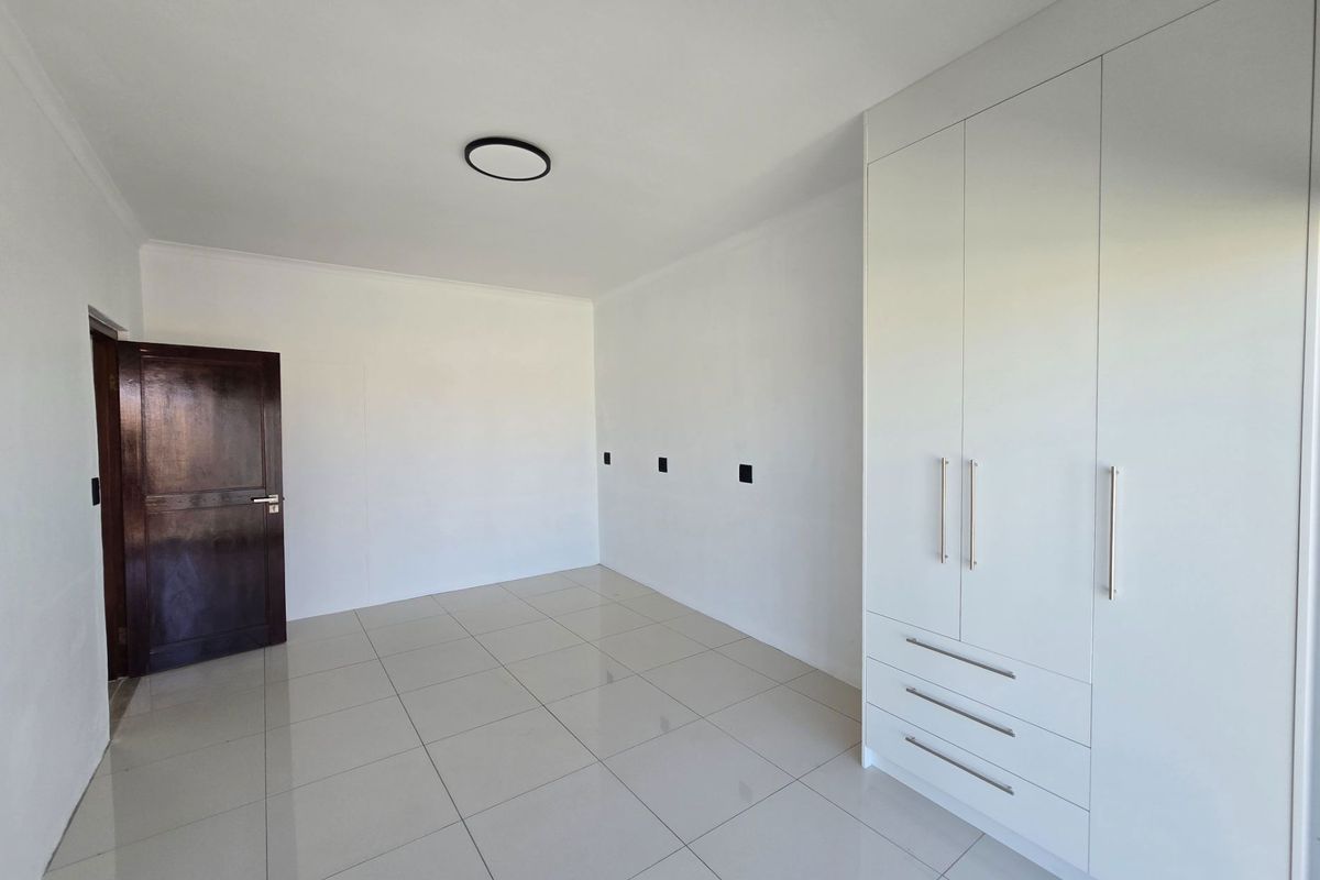 A bright, empty bedroom featuring modern white built-in cupboards, tiled flooring, and a dark wood door.