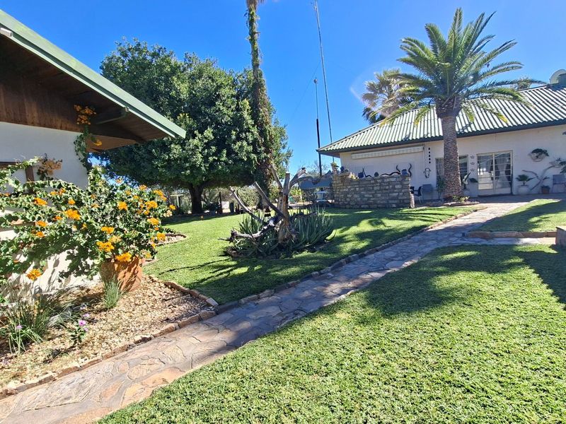 A well-maintained rear garden area featuring a stone paved walkway, lush lawn, and established landscaping between the farm building structures.