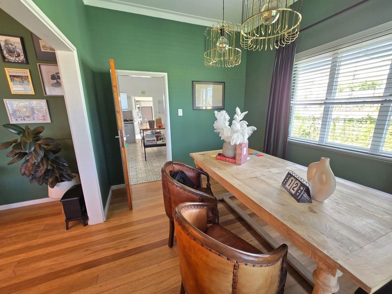 A well-maintained dining room featuring polished hardwood flooring, deep green walls, and large windows with venetian blinds, leading into an adjacent utility area.