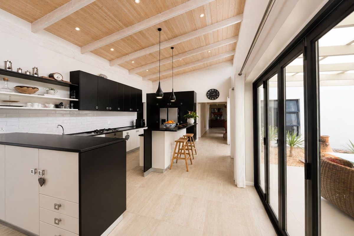 Modern kitchen featuring high-contrast cabinetry, a central island, exposed timber beam ceiling, and seamless indoor-outdoor flow through large bi-fold glass doors.