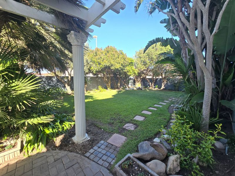 A well-maintained private garden featuring a pergola with a decorative column, a stone paver pathway, and established landscaping.