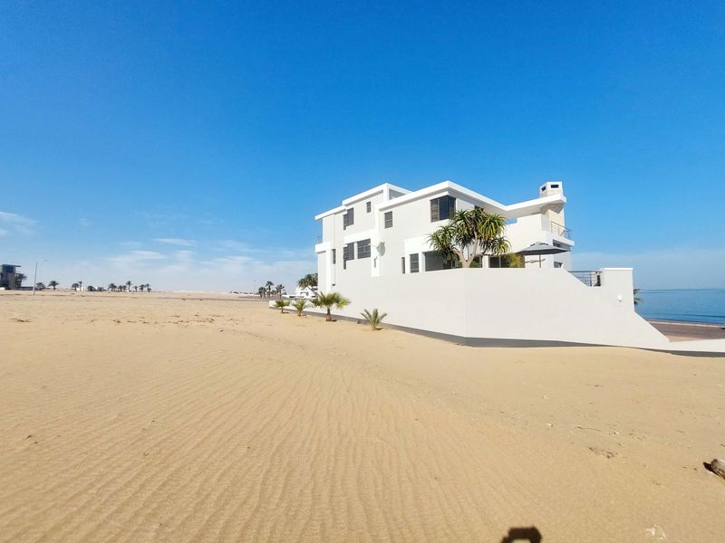 A modern multi-story beachfront house featuring a clean white facade, chimney, and balcony, situated directly on the sandy coastline.