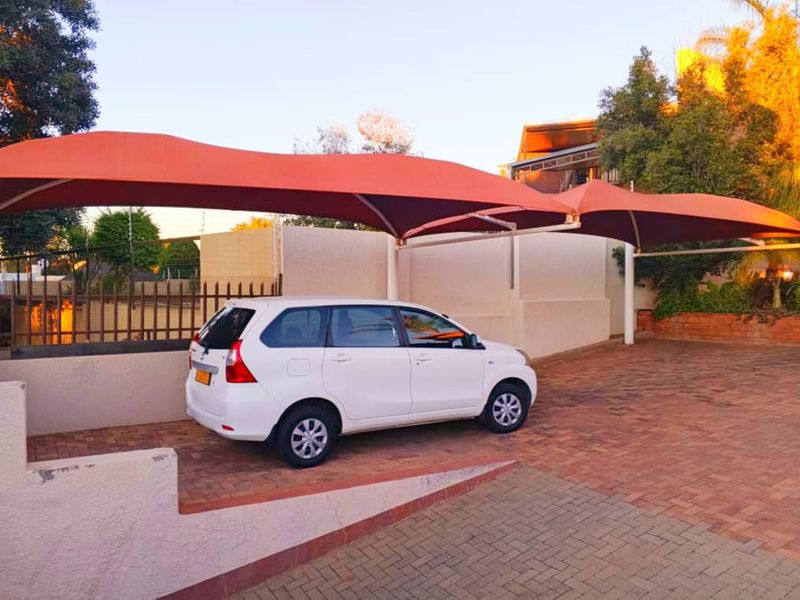 A paved driveway featuring a modern shade sail carport structure, enclosed by a perimeter wall with electric fencing.