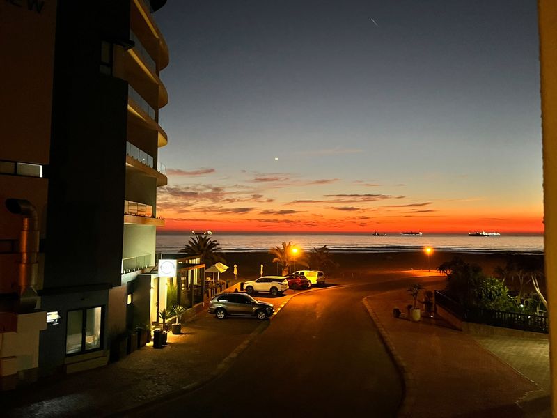 A scenic sunset view overlooking the ocean, beach, and coastal apartment building exterior at dusk.