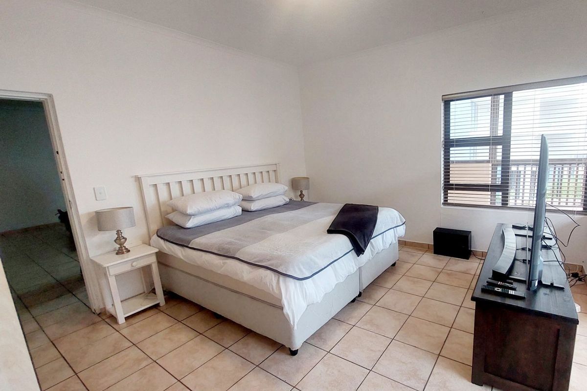 A spacious bedroom featuring neutral tiled flooring, large aluminium-framed windows with venetian blinds, and a simple ceiling light fixture.